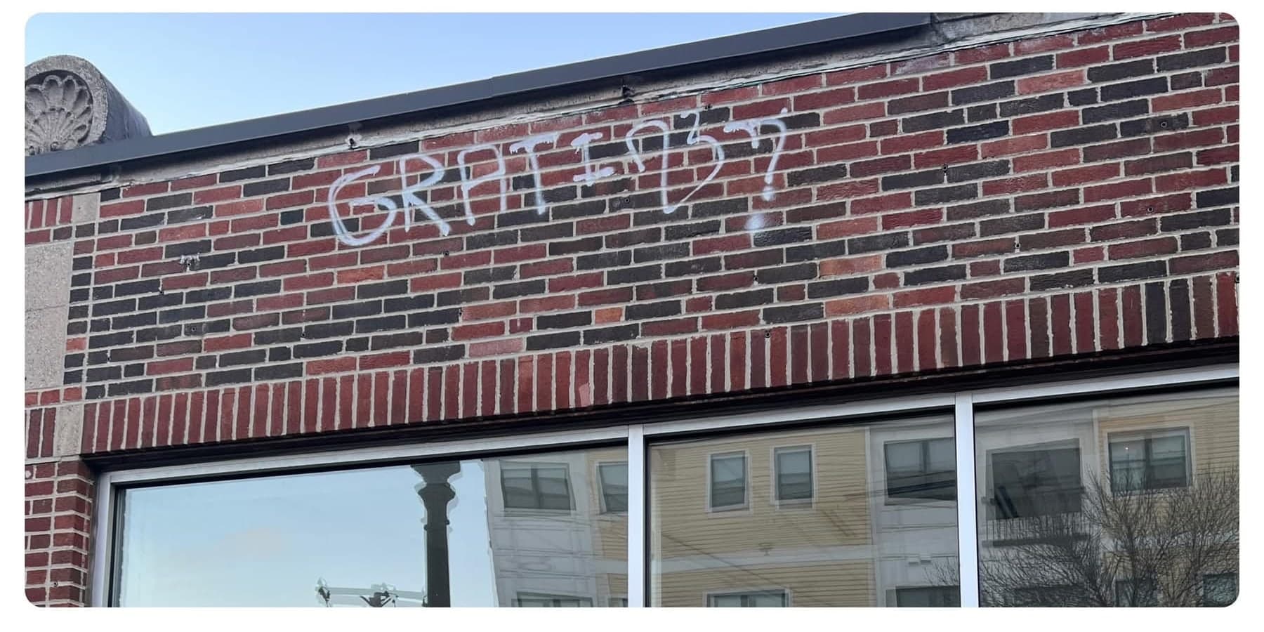 White spray-painted tag across the red brick band above a storefront window before removal