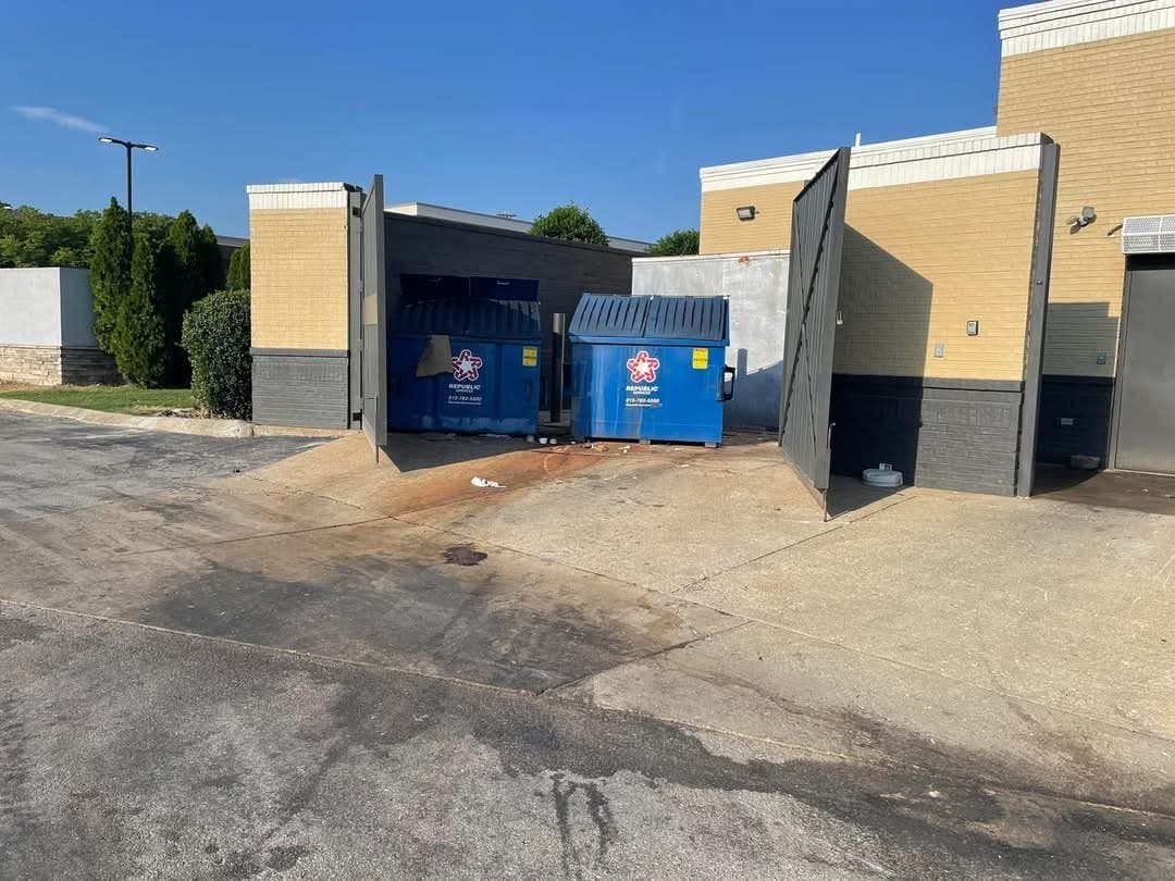 Two dumpsters inside a brick corral with its gates open, and a concrete apron in front stained dark by leachate running out toward the asphalt