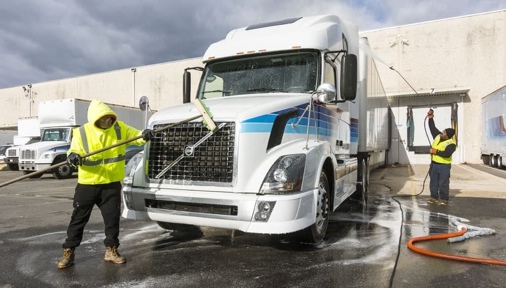 Two technicians pressure washing a white semi truck and trailer at a warehouse loading dock