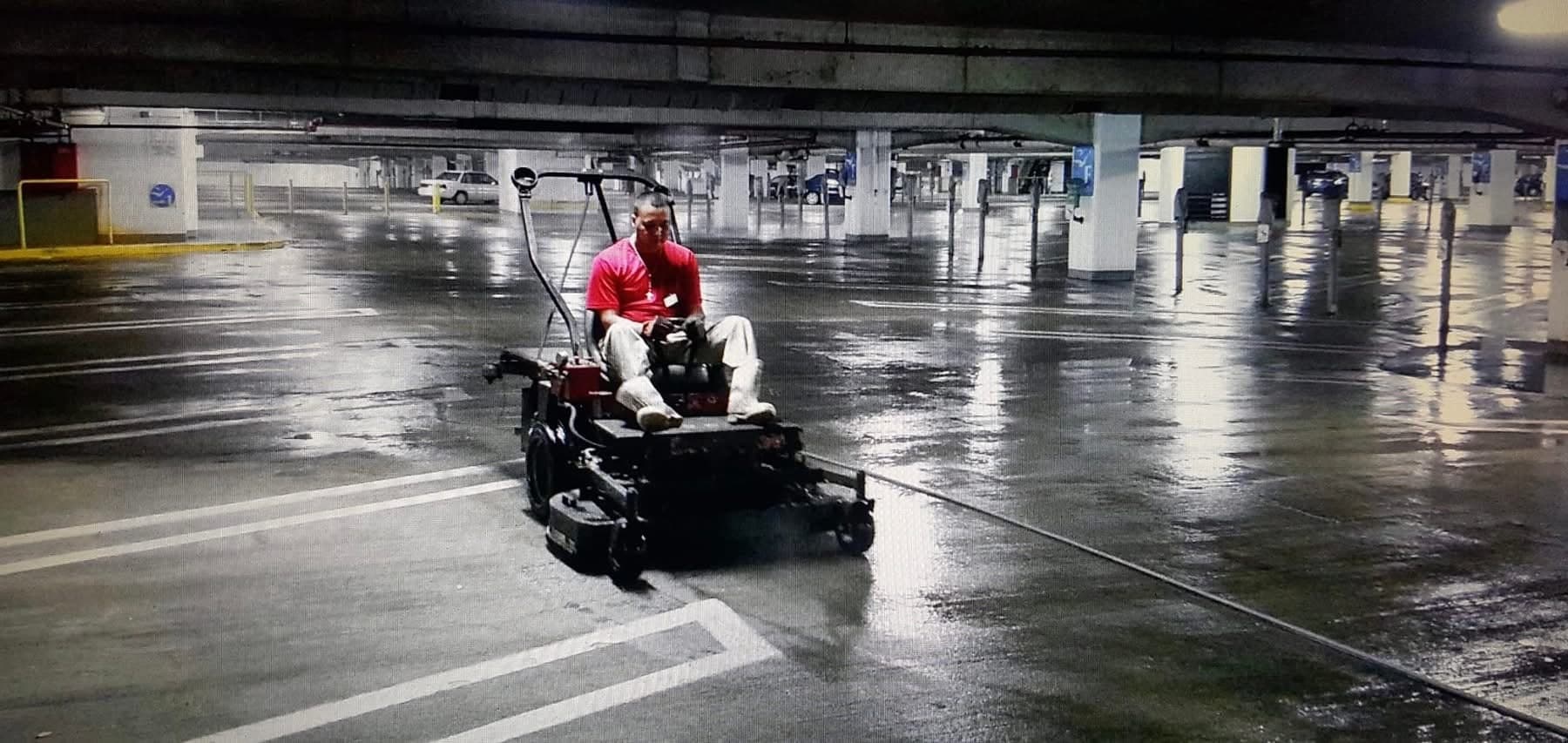 Technician cleaning a parking garage deck with a ride-on surface cleaner, wet concrete under structural columns
